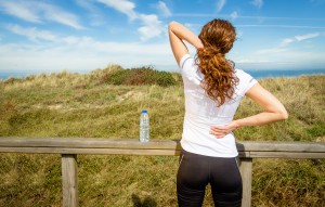 Back view of athletic young woman in sportswear touching her neck and lower back muscles by painful injury, over a nature background. Sport injuries concept.